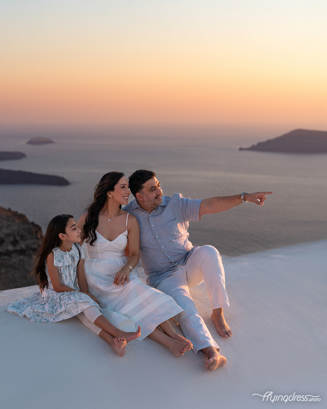 A family sits together on a rooftop in Santorini, enjoying the sunset as the father points toward the horizon. The serene backdrop of the sea and islands enhances the beauty of the moment.